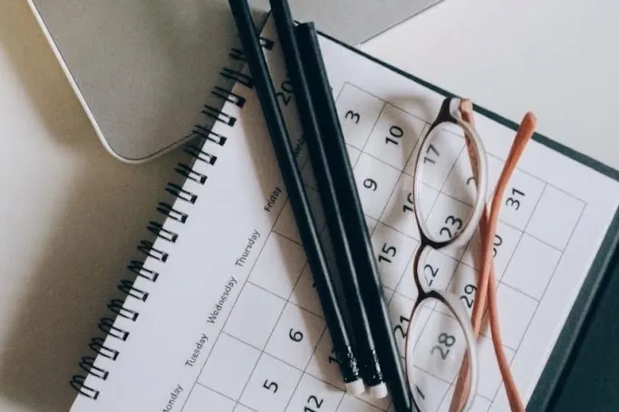 A desk calendar with two pencils and a pair of glasses on top, next to a laptop