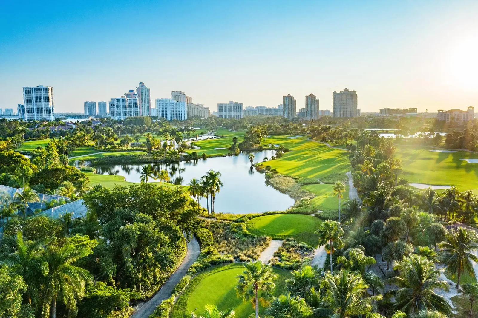 Aerial view of a golf course with lakes and palm trees, with Aventura apartment towers beyond