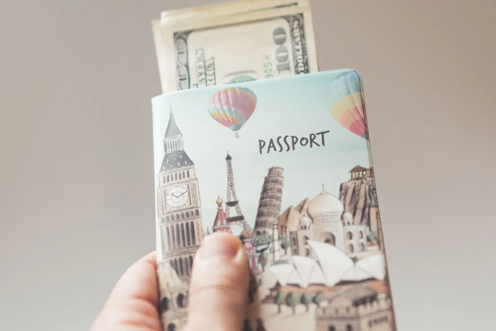 Una mano sostiene un pasaporte con funda ilustrada de monumentos del mundo, con billetes de cien dólares asomando por arriba
