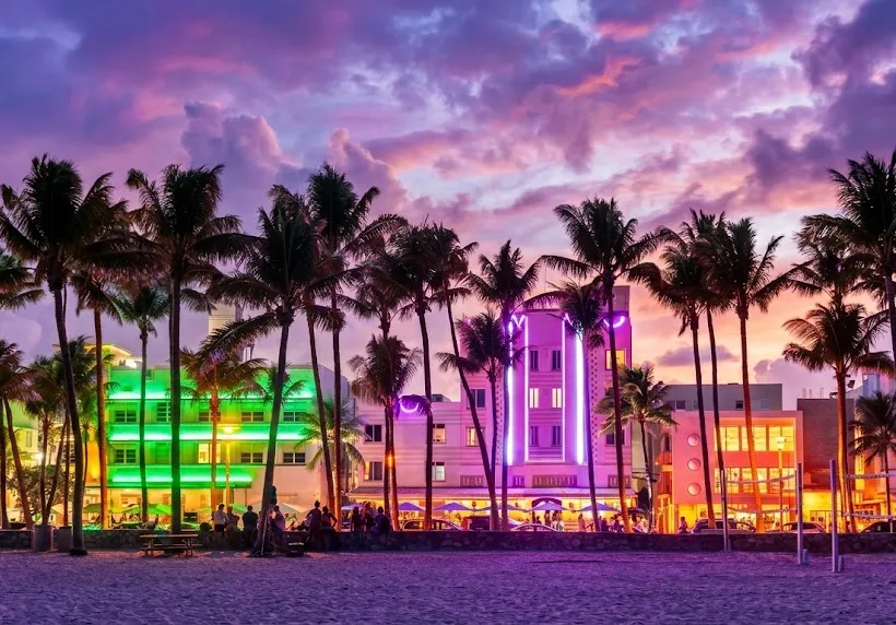 Art Deco hotels lit with colorful neon behind a row of palm trees at sunset, seen from the beach
