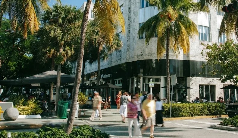Pedestrian street with palm trees, patios and people walking in front of a curved white stone building