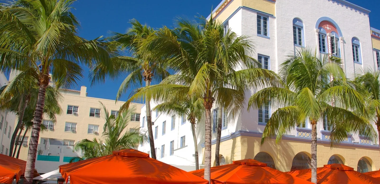 White Art Deco hotel with yellow and blue trim behind a row of palm trees and orange patio umbrellas