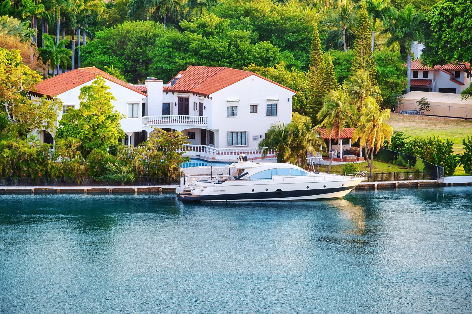 A white Mediterranean-style house with a red tile roof on a canal, with a yacht moored in front