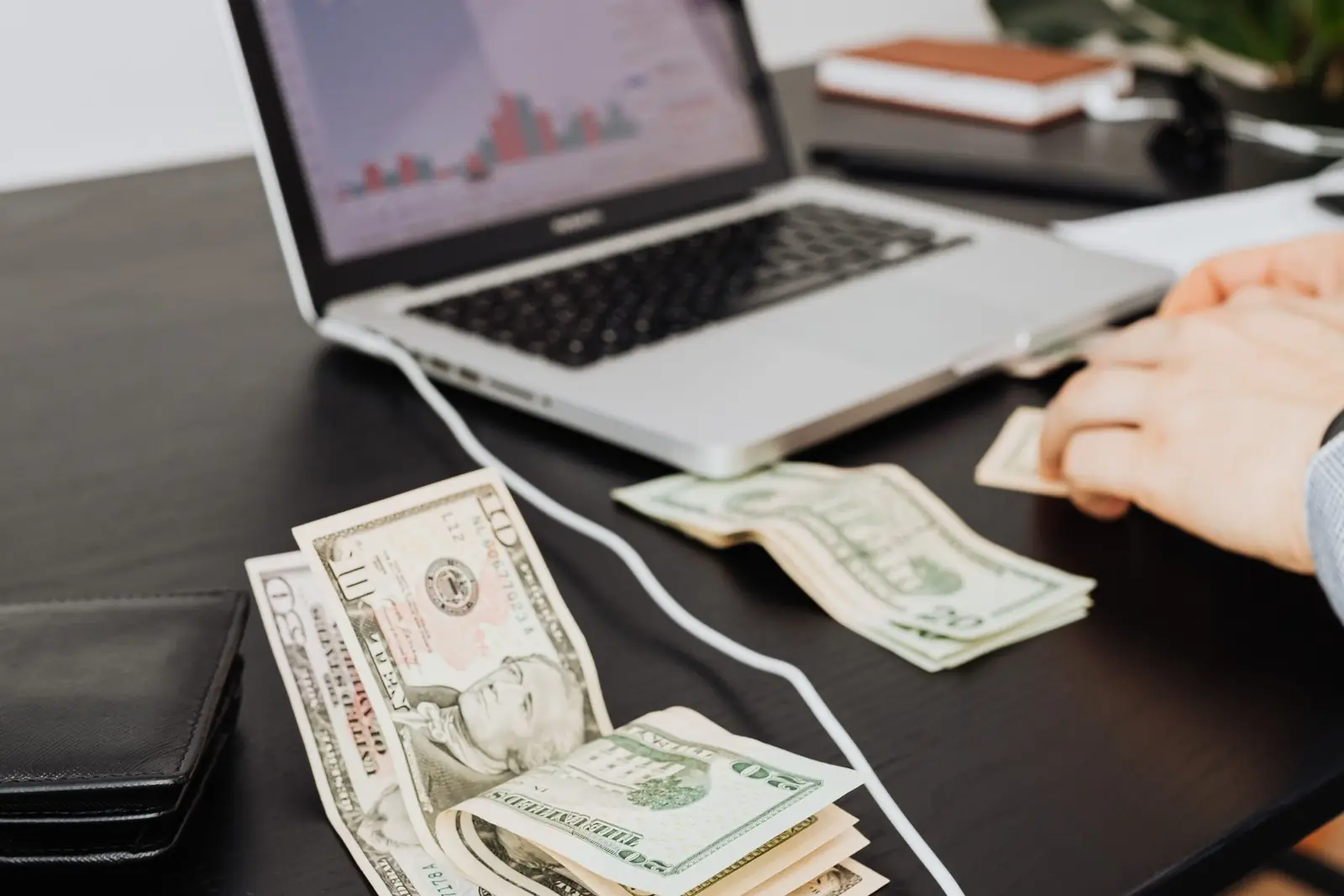 Dollar bills on a black desk next to an open laptop and a wallet