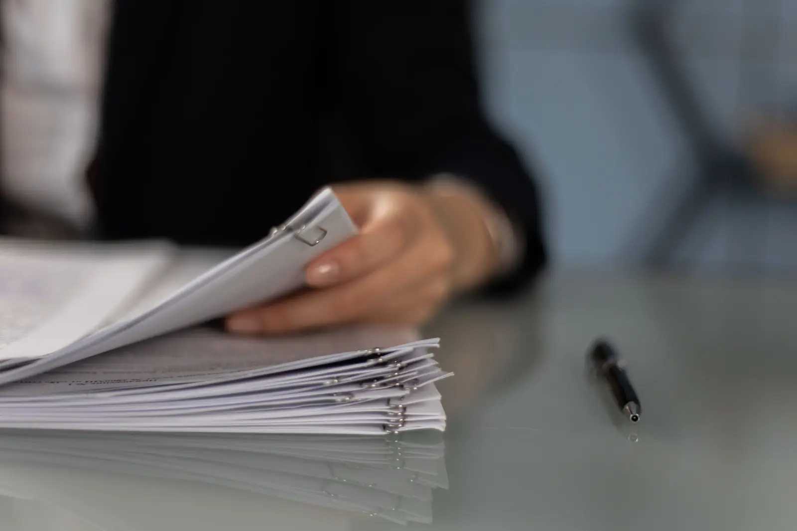 A person goes through a stack of clipped documents on a glass desk, next to a pen