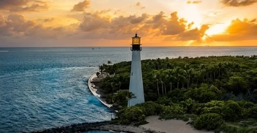 A white lighthouse by the sea among palm trees and vegetation, at sunset