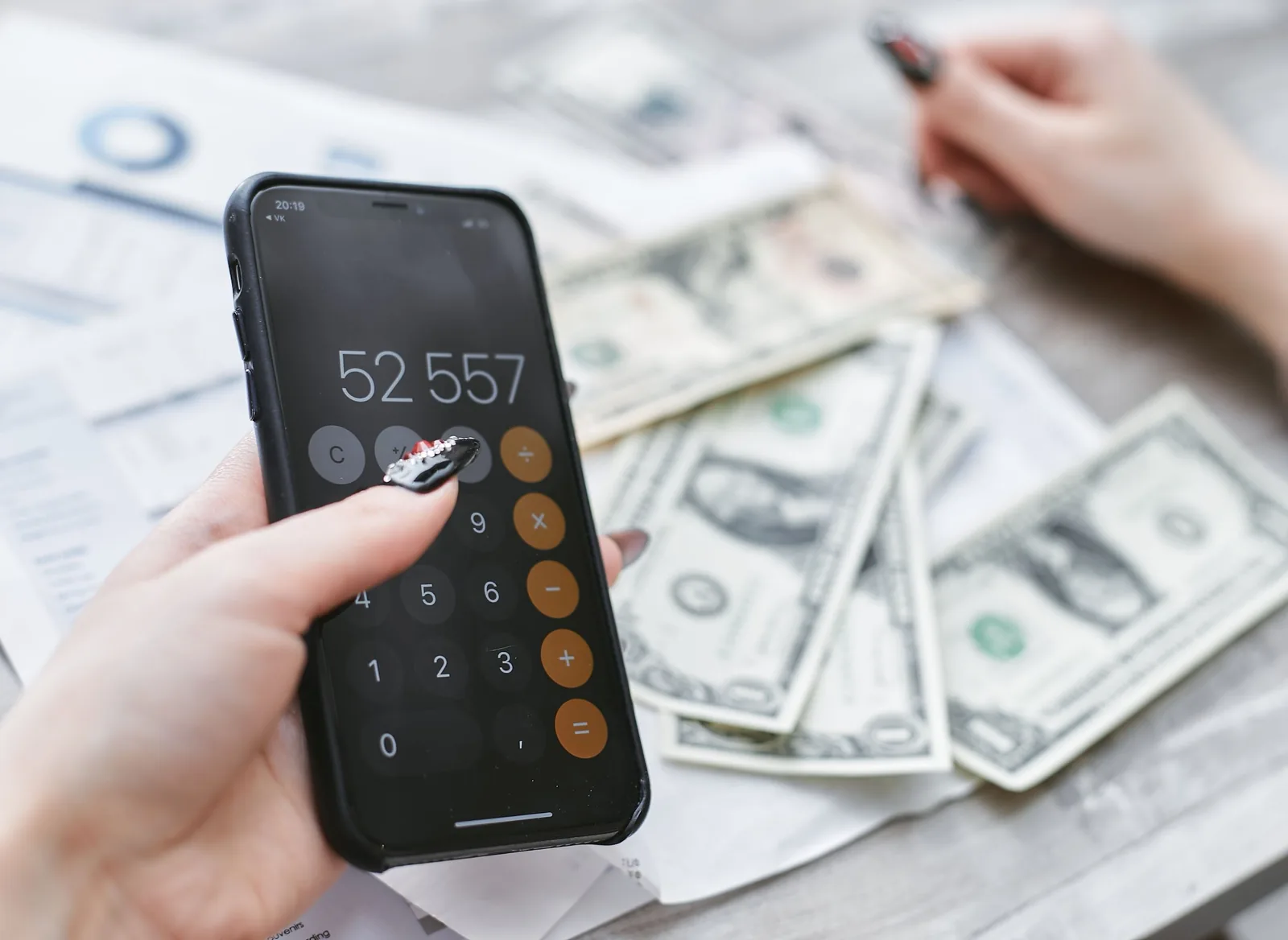 A hand holds a phone with the calculator open over dollar bills and documents