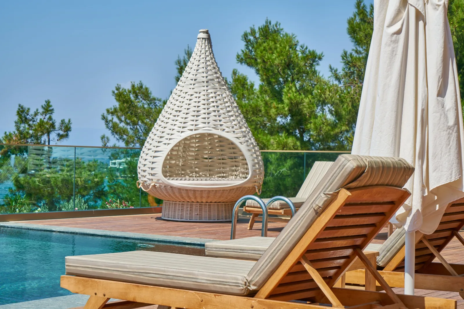 Pool terrace with wooden loungers, a white umbrella and a wicker pod chair, with trees and the sea beyond