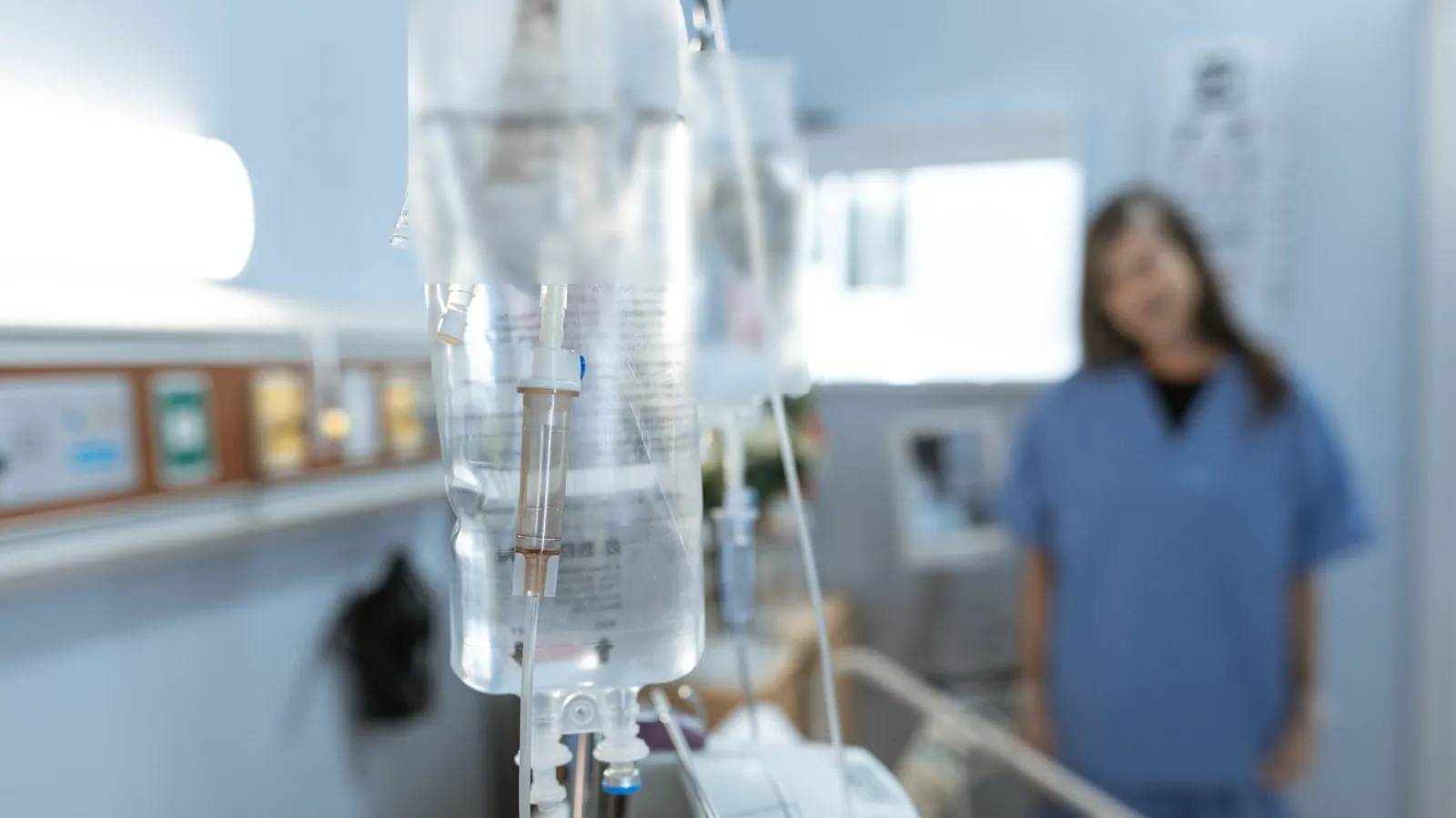 IV bags hanging beside a hospital bed, with a nurse out of focus in the background