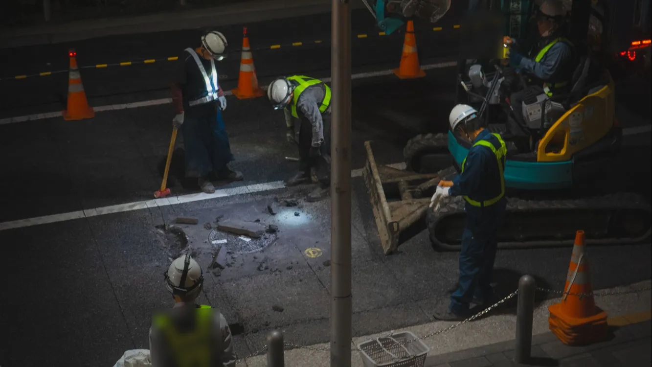 Workers in hard hats and high-visibility vests repairing a street at night beside an excavator