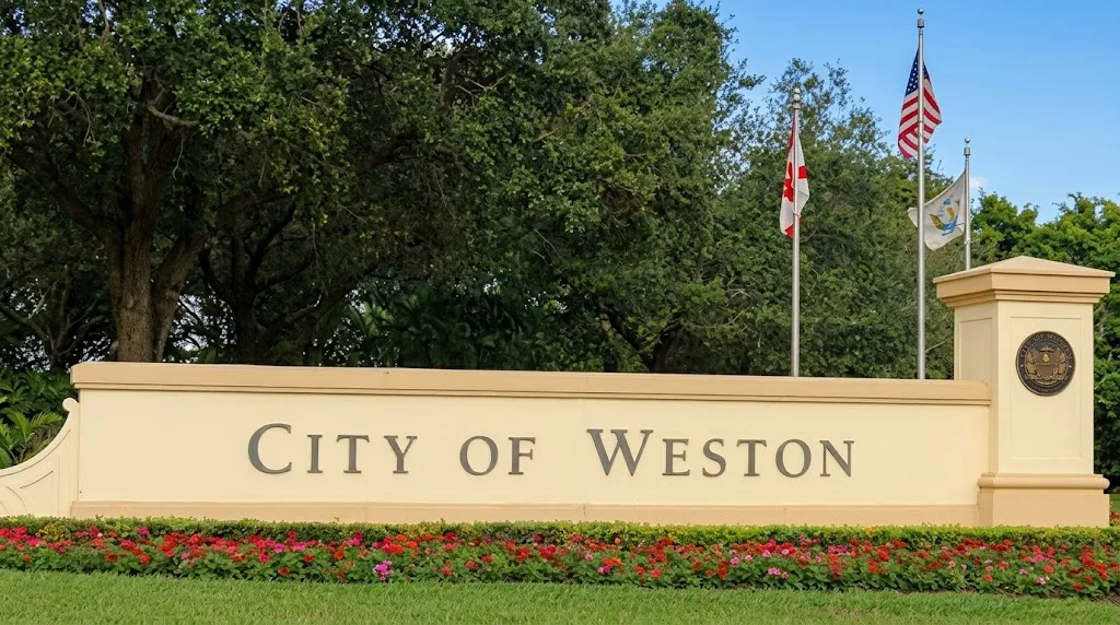 "City of Weston" welcome sign on a cream wall with red and pink flowers in front, flags and trees behind