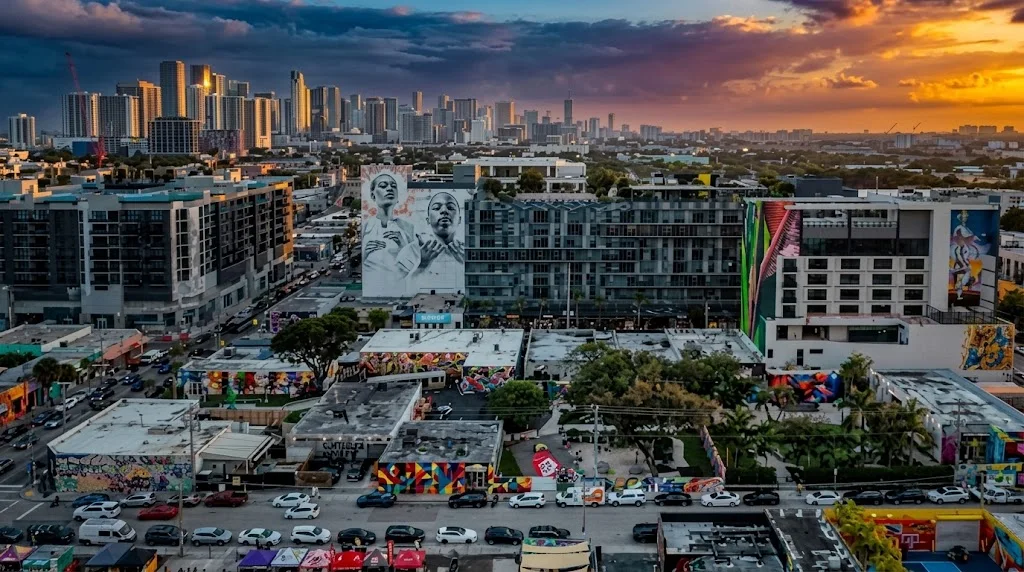 Aerial view of Wynwood at sunset: warehouses and buildings covered in colorful murals, with the downtown Miami skyline behind